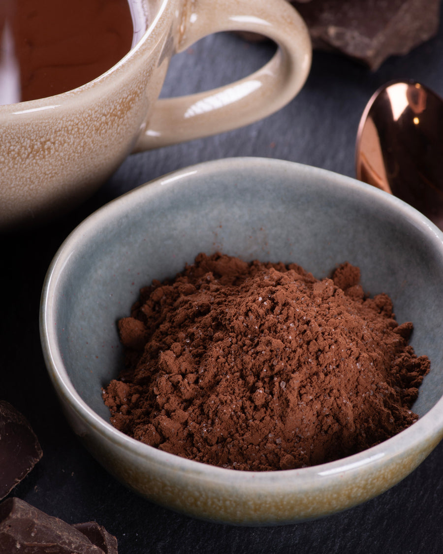 Cocoa powder in a bowl with a mug and chocolate pieces on a dark surface