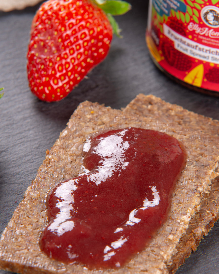 Cracker with strawberry jam on a dark surface, next to a strawberry and a jar of strawberry jam.