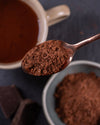 Cocoa powder on a spoon with a cup of hot chocolate and cocoa blocks in the background.