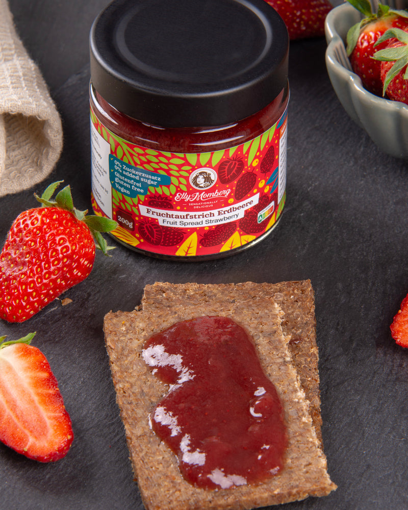 Jar of strawberry jam with a spread on a cracker, surrounded by strawberries on a dark surface.