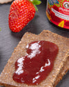 Cracker with strawberry jam on a dark surface, next to a strawberry and a jar of strawberry jam.