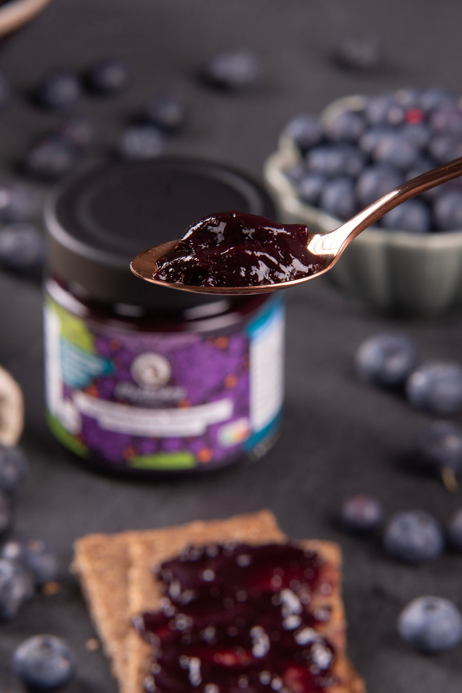 Jar of blueberry jam with a spread on crackers, surrounded by blueberries on a dark surface