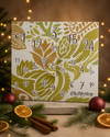 Veganer Adventskalender Deluxe 2026 als Weihnachtsgeschenk mit Tannengrün, Burgunderkugeln und warmem Licht