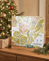 Veganer Adventskalender Deluxe 2026 mit 24 Überraschungen und 1500g vor beleuchtetem Weihnachtsbaum