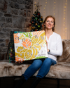 Frau präsentiert den veganen Adventskalender Deluxe 2026 mit 1500g vor Weihnachtsbaum und Lichterkette